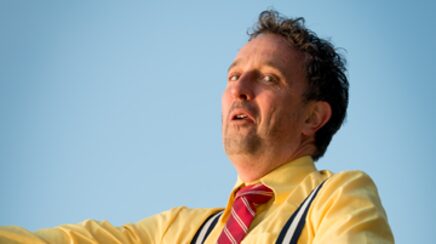 Marcel Harmsen mid-gesture in a yellow shirt and striped tie against open sky